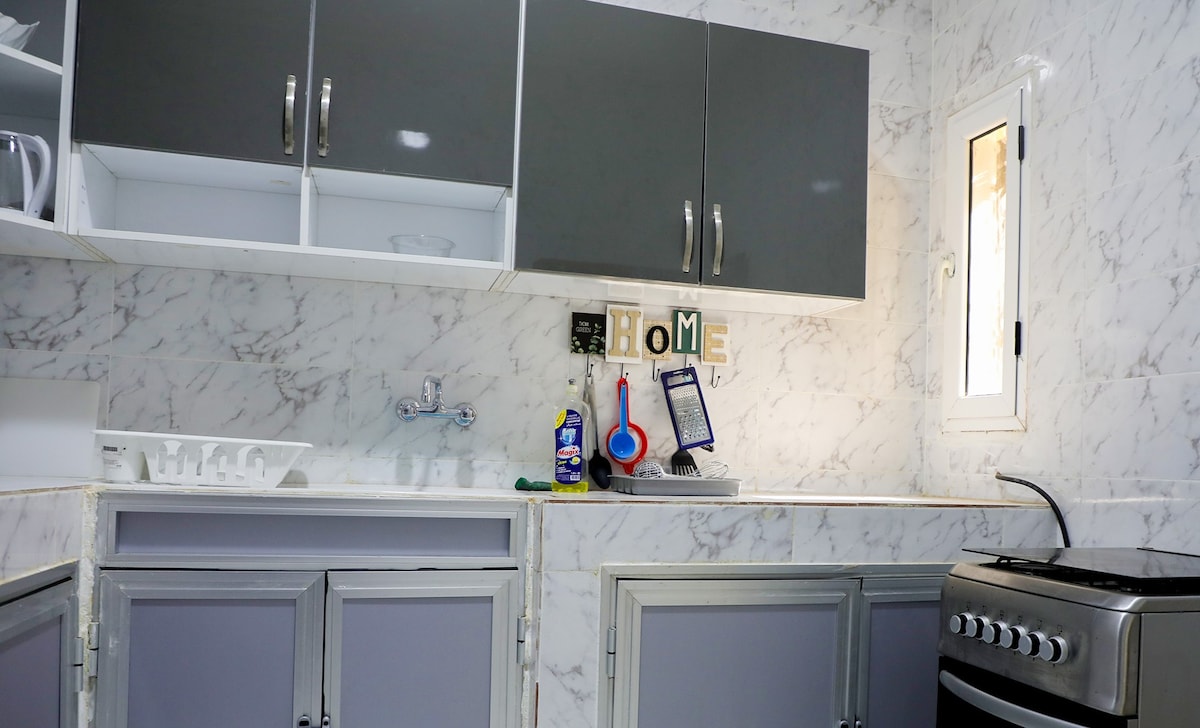 A modern kitchen features sleek grey cabinets with white accents. A marble countertop runs along the wall, showcasing kitchen essentials and utensils. A stove is positioned to the right, while a small window allows natural light to illuminate the space.