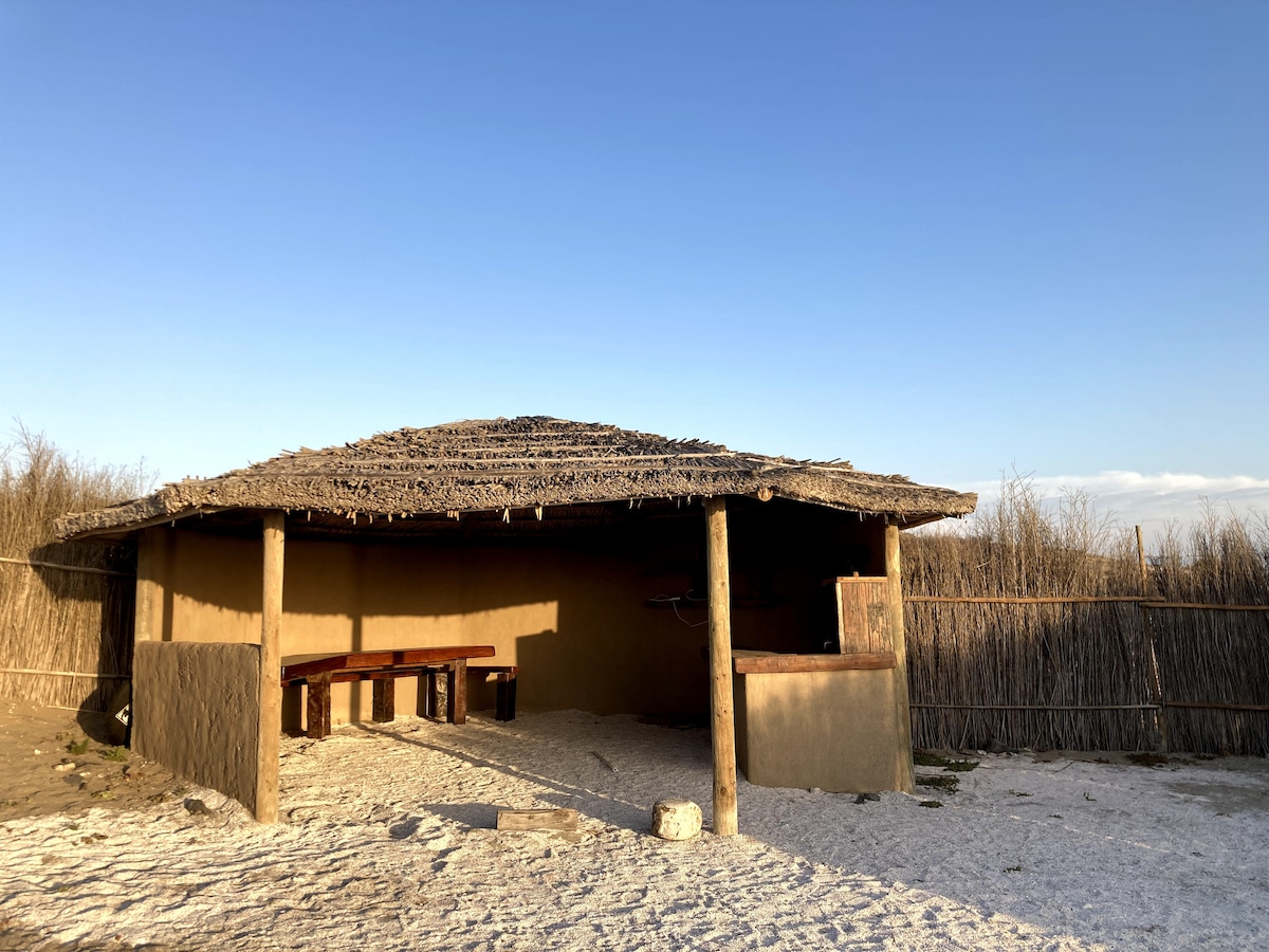 A rustic structure is situated under a clear blue sky, featuring a thatched roof supported by wooden poles. An open area in front displays a sandy floor, with long benches arranged along one side and a simple cooking space visible within the structure.