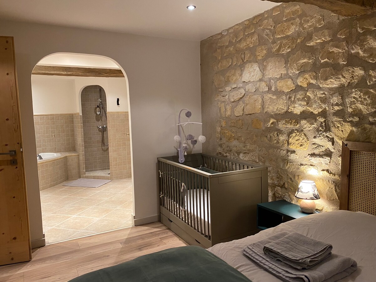 A cozy bedroom area is visible, featuring a spacious bed and a baby cot positioned nearby. A doorway leads to a large bathroom, showcasing a combination of modern tile and stone materials. Soft lighting illuminates the room, creating a warm and calming ambiance.