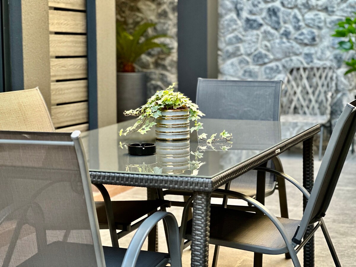 An outdoor dining area features a glass-top table surrounded by four metal-framed chairs, offering a blend of comfort and style. A small potted plant rests at the center, complemented by a stone wall in the background, enhancing the serene outdoor setting.