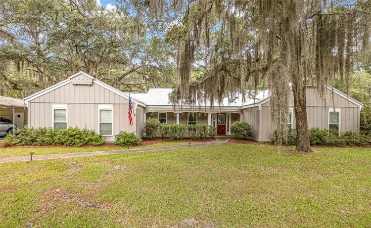 A single-story house is set amid lush greenery, featuring a light grey facade and a red front door. Spanish moss hangs from nearby trees, and the neatly maintained lawn adds to the welcoming appearance of the property.