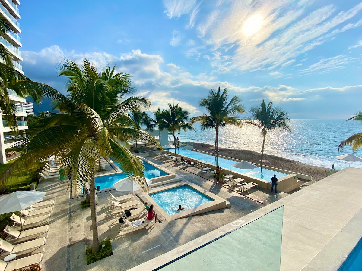 Multiple swimming pools are surrounded by palm trees, with chaise lounges positioned nearby. The ocean is visible in the background, and the sun is positioned low in the sky, casting a warm glow over the landscape.