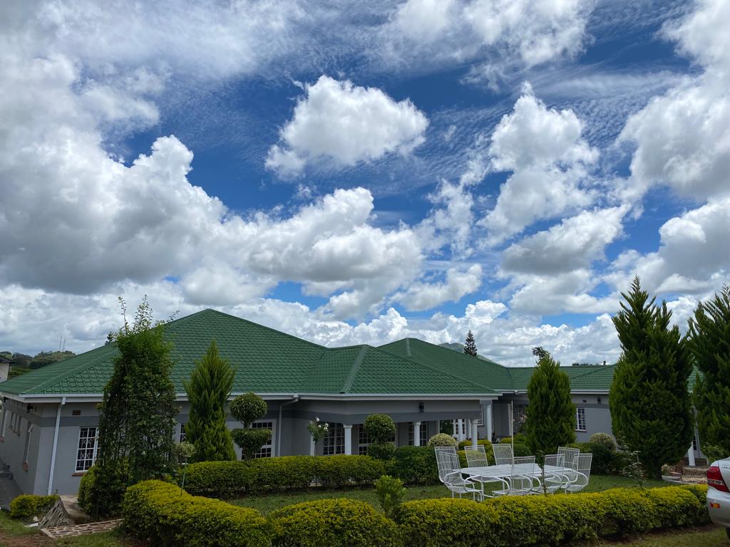 The exterior of the property features a well-maintained garden with neatly trimmed hedges and shrubs, framed by trees. A spacious green roof complements the building, while a blue sky with scattered clouds provides a serene backdrop. Outdoor seating is arranged on a patio area.