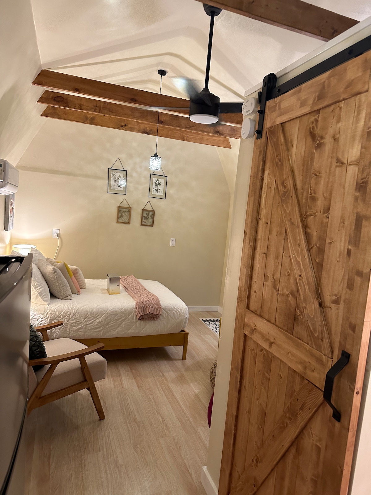 A cozy bedroom is viewed from the entrance, featuring a spacious bed with decorative pillows and a soft blanket. Exposed wooden beams create a warm ambiance. A sliding barn door adds rustic charm, while subtle lighting fixtures enhance the inviting space.