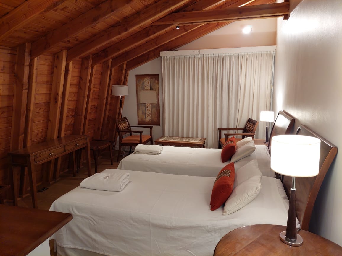 A cozy bedroom features two spacious beds, each topped with white linens and accent pillows. Natural wood beams adorn the ceiling, while warm lighting from bedside lamps creates a calming effect. A writing desk and chairs provide additional comfort, with a large window covered by light curtains.