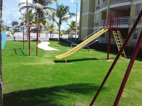 An outdoor play area is highlighted by a yellow slide and two red swings, set on a grassy lawn. Palm trees surround the space, contributing to a relaxed environment. The nearby building offers balconies that overlook the play area.