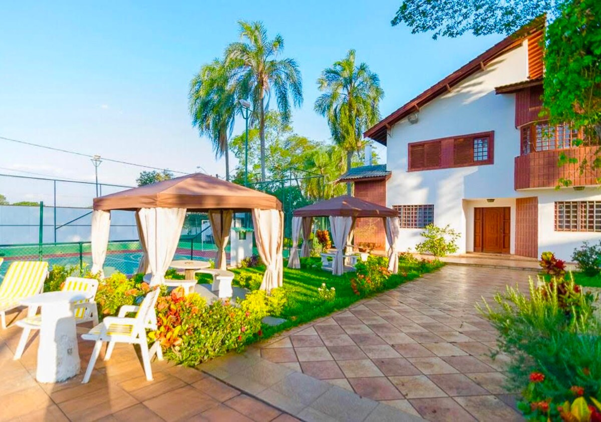The outdoor area displays well-maintained gardens with blooming flowers and lush greenery. Shaded seating areas are provided by canopies, along with white tables and chairs. A tennis court is visible in the background, and the exterior of the house features wooden shutters, harmonizing with the serene environment.