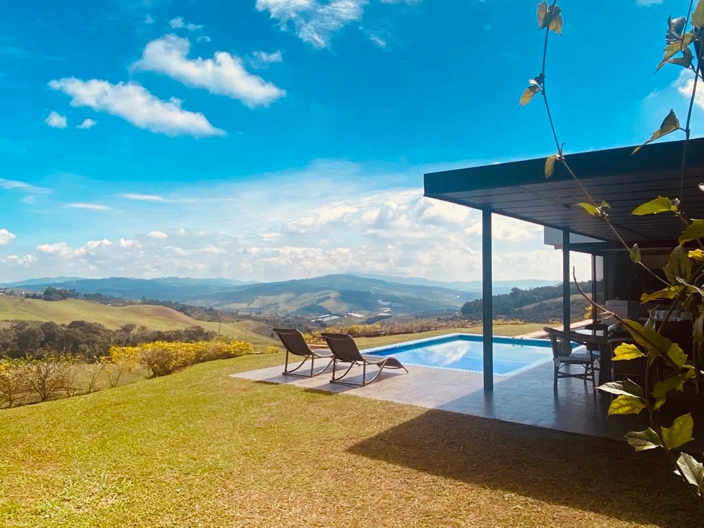 An outdoor area is presented featuring a clear blue sky over rolling hills. A pool reflects the scenery and is accompanied by two lounge chairs on a grassy lawn. Dining space is visible under a roof, seamlessly blending with the tranquil surroundings.