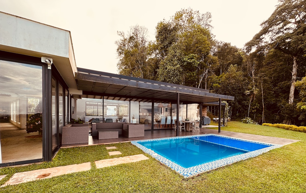 The exterior of the villa showcases a patio area with modern outdoor furniture, overlooking a private pool. Lush greenery surrounds the property, while large windows provide views of the natural landscape. The pool is framed with decorative tiles, enhancing the outdoor space.