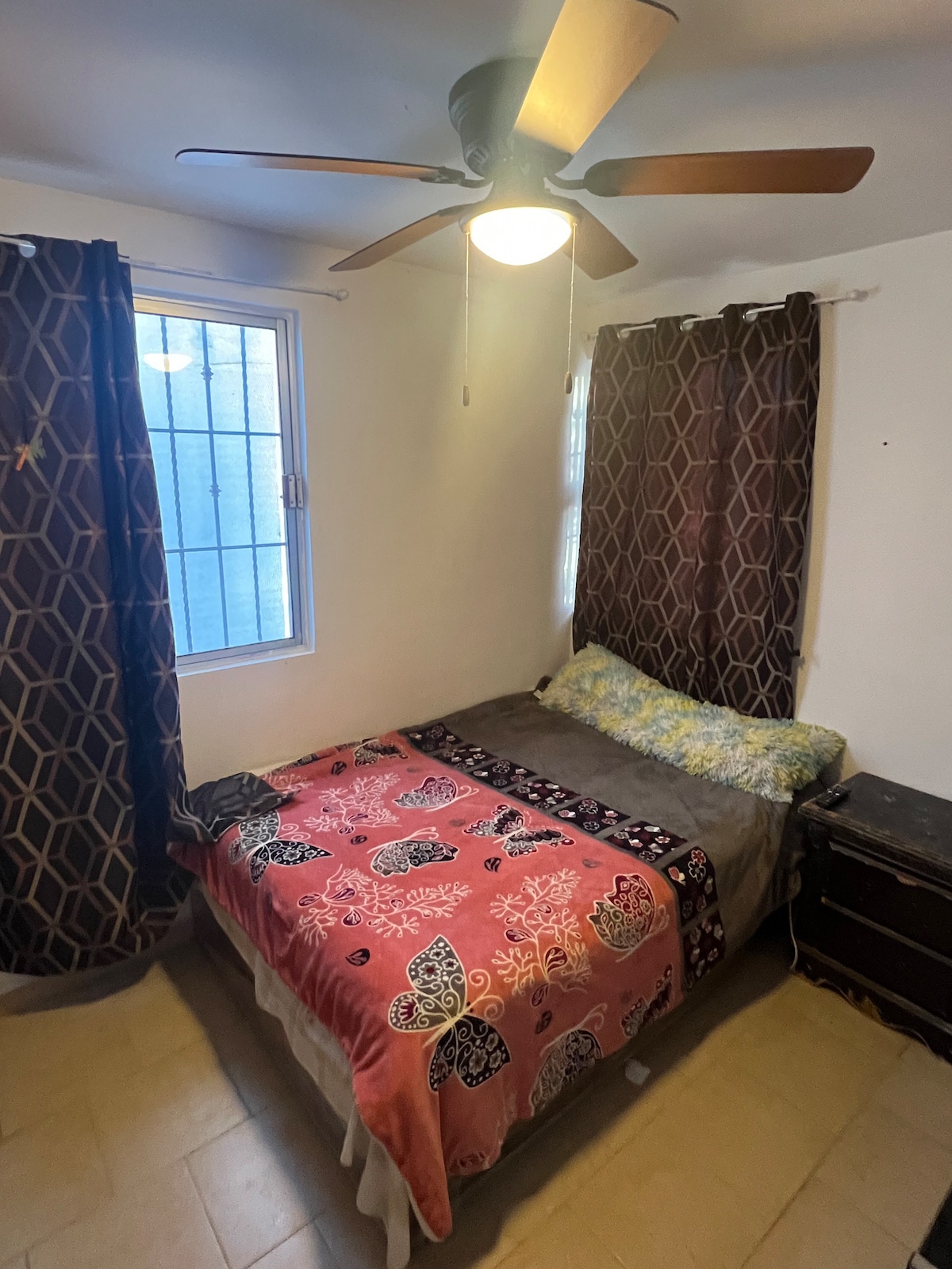 A cozy bedroom features a spacious bed covered with a patterned blanket. Natural light enters through a window adorned with a sheer curtain. A ceiling fan provides ventilation, while dark curtains offer privacy. A small nightstand is positioned beside the bed.