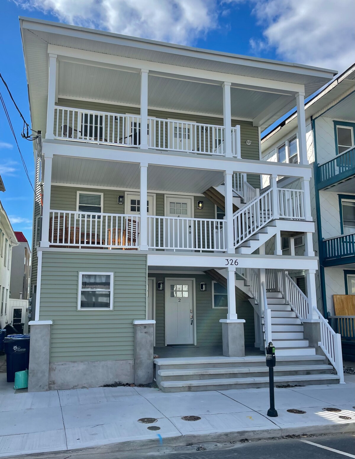 The exterior of a multi-story condominium building is showcased, featuring a pale green facade and white trim. Multiple balconies and a staircase create visual interest, while the entrance door is framed by lit sconces on either side. Clear blue skies serve as a backdrop.