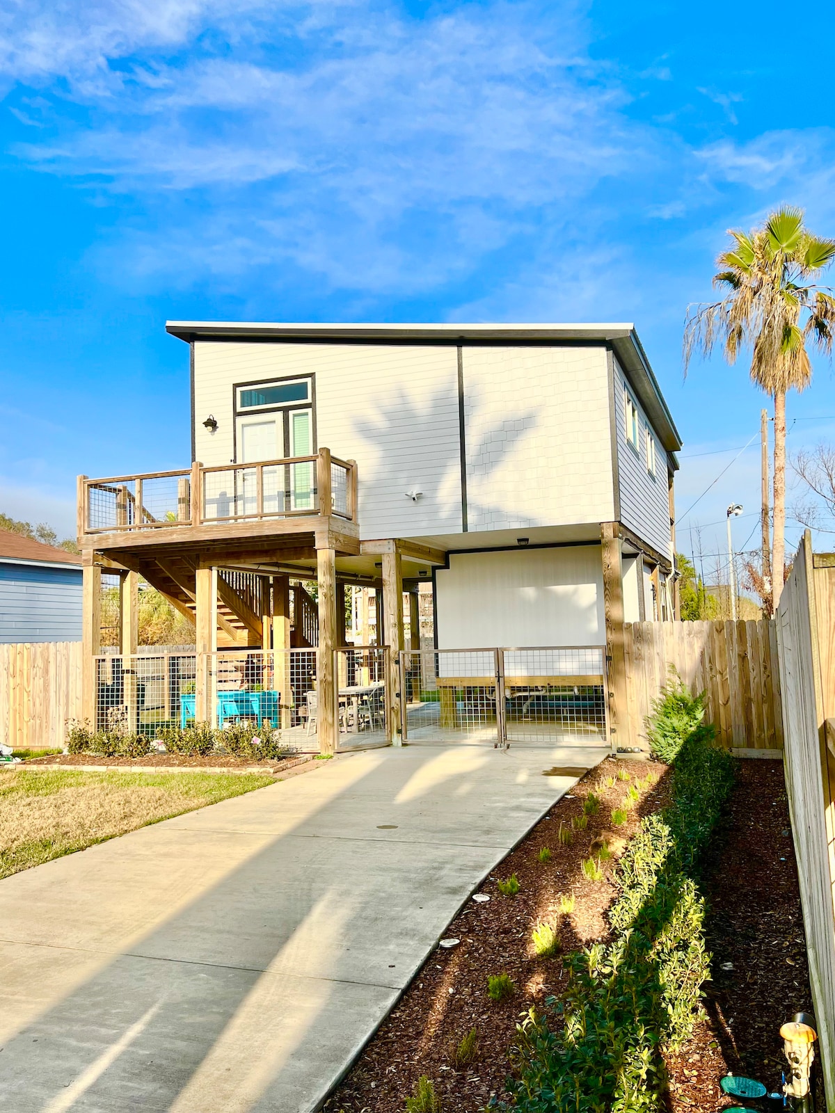 A two-story unit is shown, set against a clear blue sky. The exterior features a light-colored facade with large windows. A deck is visible above, and a fenced yard surrounds the property, framed by well-maintained landscaping and palm trees.