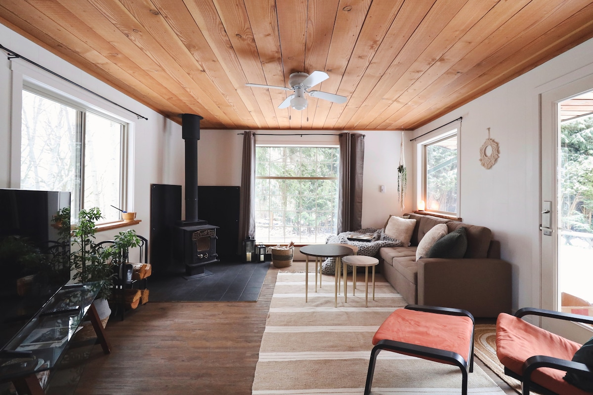 The living room features a woodstove alongside a couch that converts into a double bed. Two armchairs sit on a neutral-toned rug, and a coffee table set is positioned centrally. Large windows allow natural light to fill the room, while a ceiling fan enhances airflow.