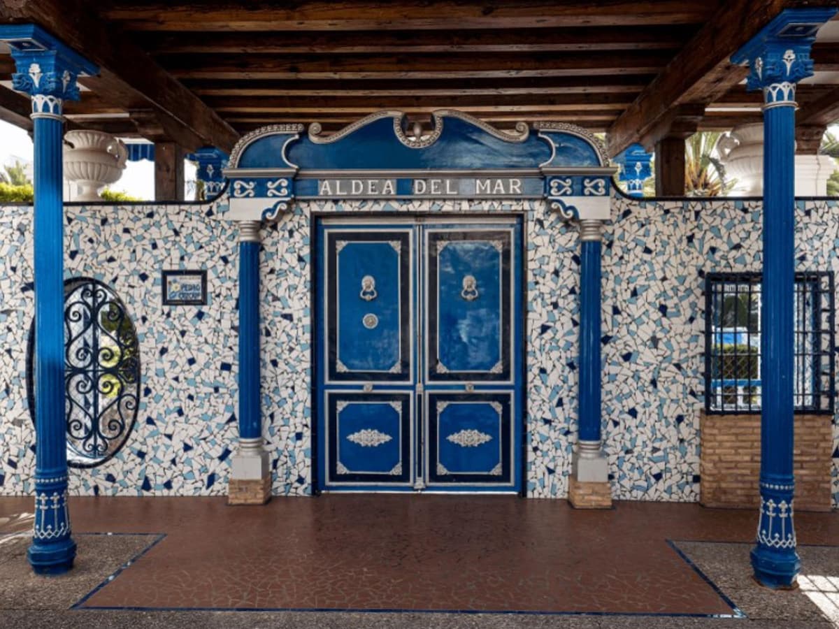 The entrance features a striking blue and white façade, characteristic of Andalusian style. Decorative tiles adorn the wall, complemented by dark wooden beams overhead. Two ornate blue doors stand at the center, framed by intricately designed columns that enhance the welcoming presence of the building.