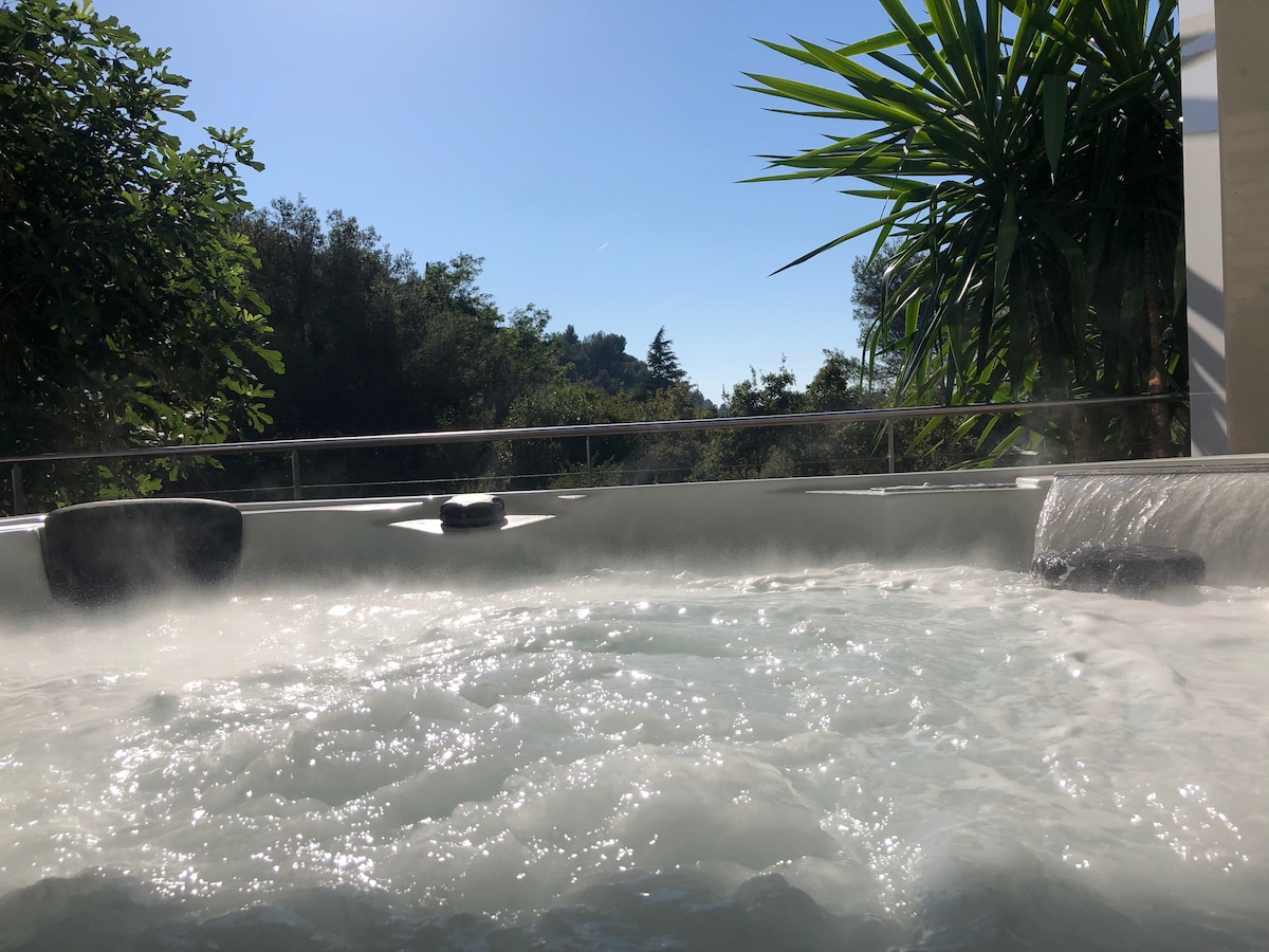 A bubbling jacuzzi is situated outdoors, surrounded by lush greenery. The clear sky above allows sunlight to reflect on the water's surface, creating a serene atmosphere. The edge of the jacuzzi is framed by tropical plants, enhancing the private setting.