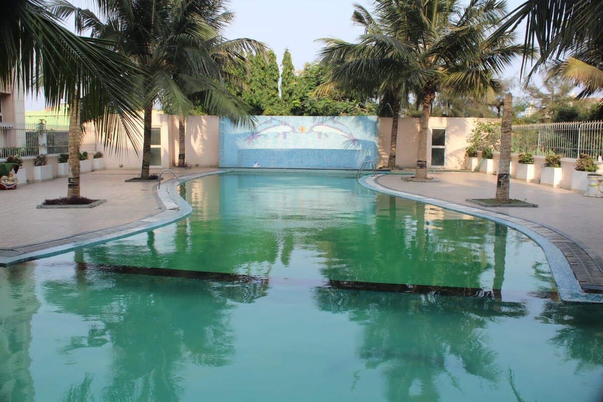 A large swimming pool is surrounded by neatly arranged palm trees, creating a serene environment. A mural decorates the backdrop of the pool area, while tile borders outline the pool's shape. Lush greenery is visible beyond the pool area, enhancing the space.