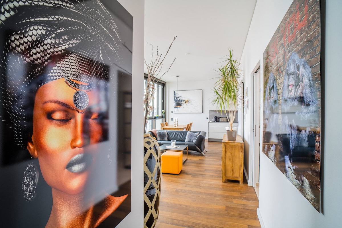 The hallway features a striking artwork on the left, leading into a bright living space with comfortable seating and natural light. A potted plant adds a touch of greenery, while a blend of modern décor and wooden accents enhances the inviting atmosphere.