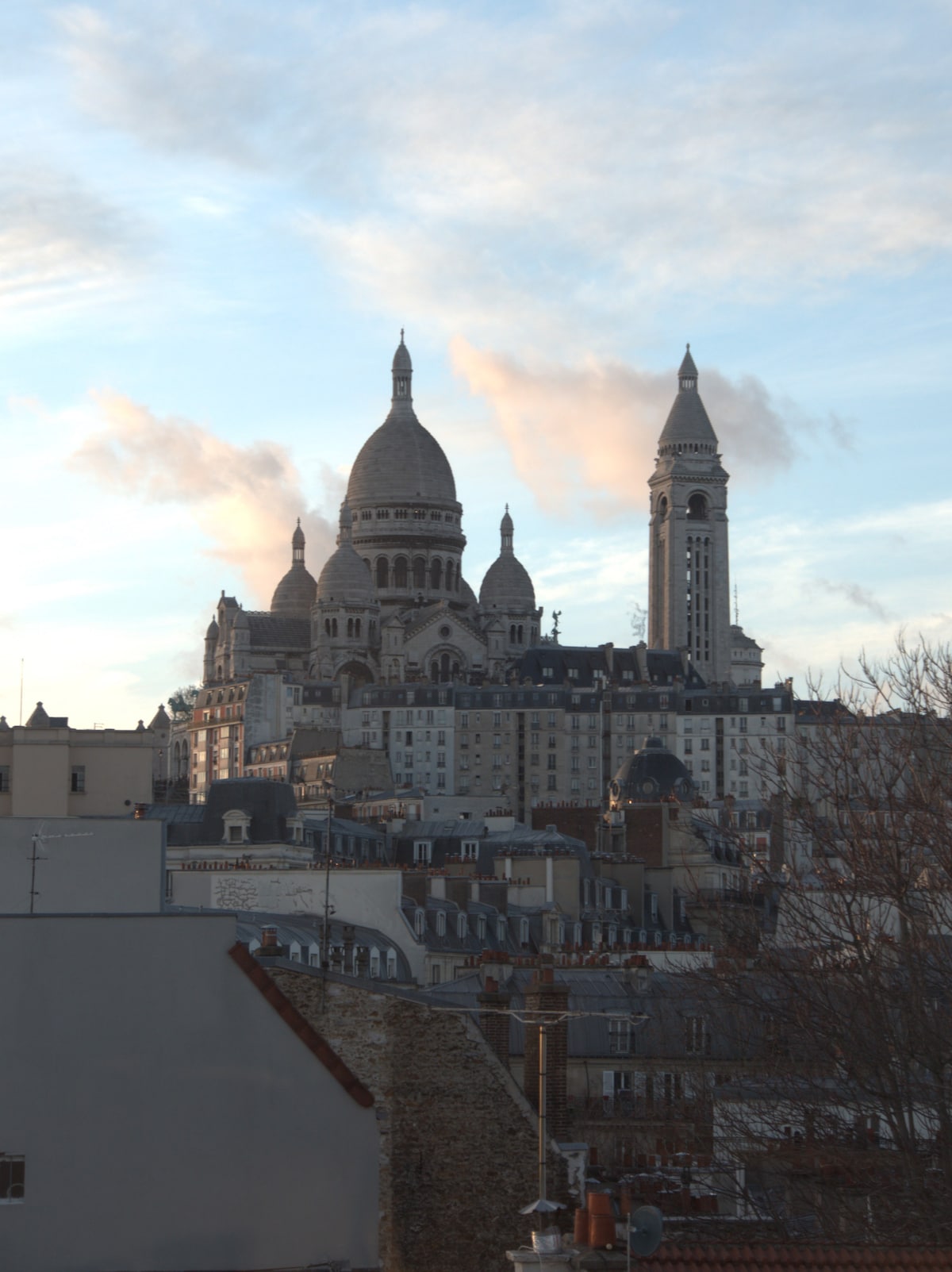 The image captures a distant view of the Sacré-Cœur Basilica, showcasing its distinctive dome and bell towers rising above the surrounding rooftops. Soft clouds in a pastel sky provide a serene backdrop to this iconic landmark, highlighting its architectural details.
