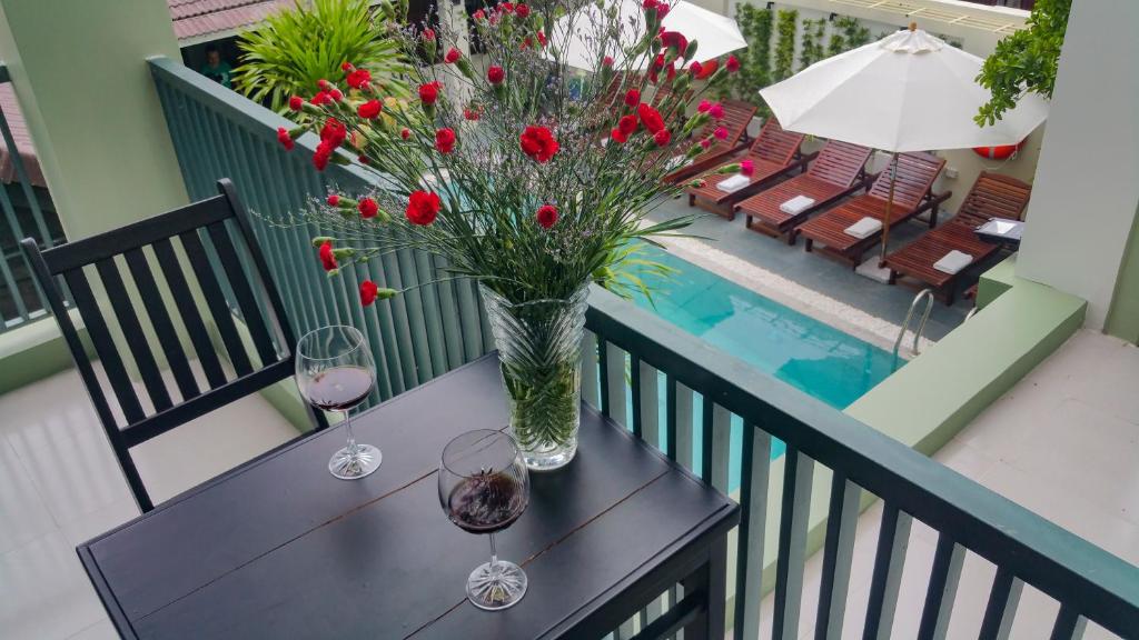 A small outdoor table is set with two glasses of wine, accompanied by a vase filled with red flowers. In the background, a swimming pool is framed by lounge chairs and umbrellas, while lush greenery surrounds the area.