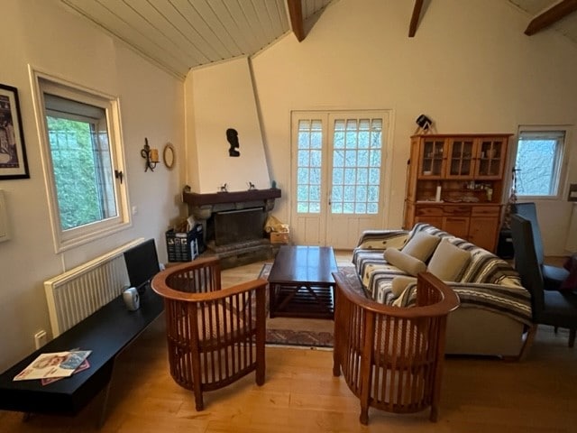 The living area features a functional fireplace with wood provided, accompanied by comfortable seating arrangements. A central coffee table is situated between two armchairs, while natural light enters through several windows, illuminating the wooden floors and adding warmth to the space.