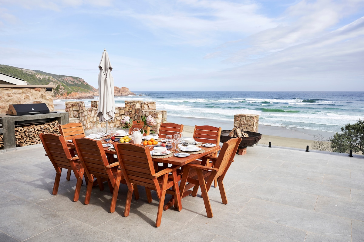 An outdoor dining area features a large wooden table surrounded by eight matching chairs, set against a backdrop of the ocean. A large umbrella provides shade, and the coastline can be seen in the distance. The patio is designed with stone walls enhancing the seaside ambiance.