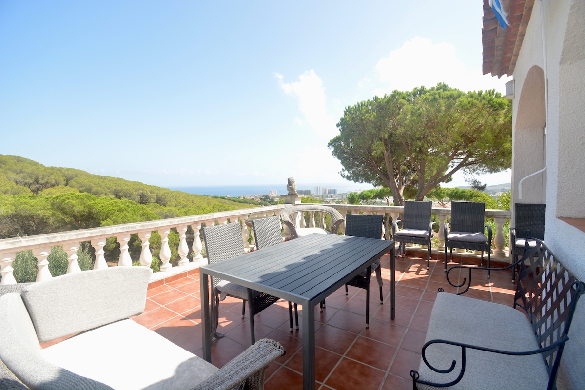 A spacious terrace features a large dining table surrounded by comfortable chairs. Views of the sea and surrounding greenery are visible, with the sky bright and clear. The terrace is partially shaded by a large tree, enhancing the outdoor space.