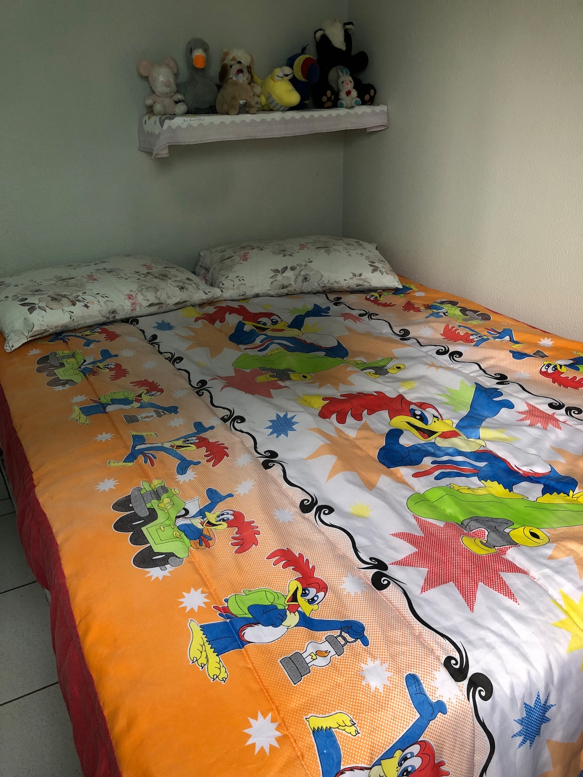 A spacious bed is dressed with a colorful comforter featuring cartoon characters. Two pillows rest at the head of the bed. Above, a shelf displays a collection of plush toys, adding a playful touch to the sleeping area.
