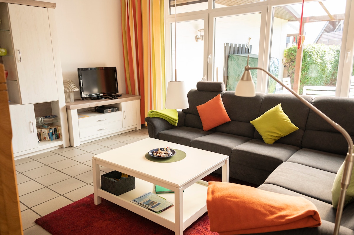 A modern living room features a gray sectional sofa adorned with bright orange and green cushions. A white coffee table occupies the center, accompanied by a decorative tray. Soft natural light filters through large glass doors, complementing the fresh color palette of the decor.