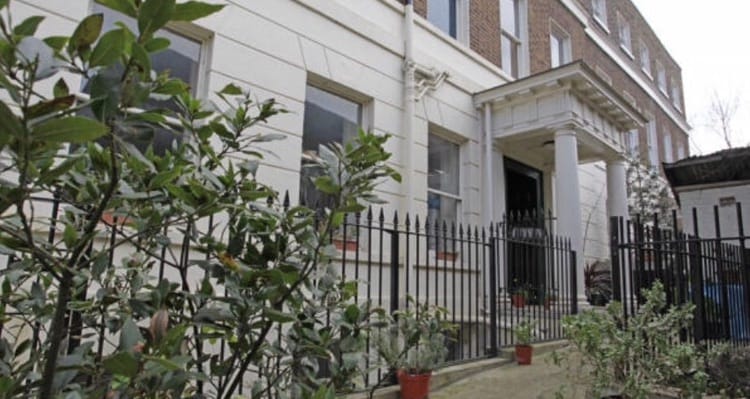 The exterior of a Grade II Listed Georgian townhouse is showcased, highlighting its elegant white facade and tall windows. A low black fence encloses a garden area with potted plants. The entrance features a prominent portico, providing a welcoming access point.