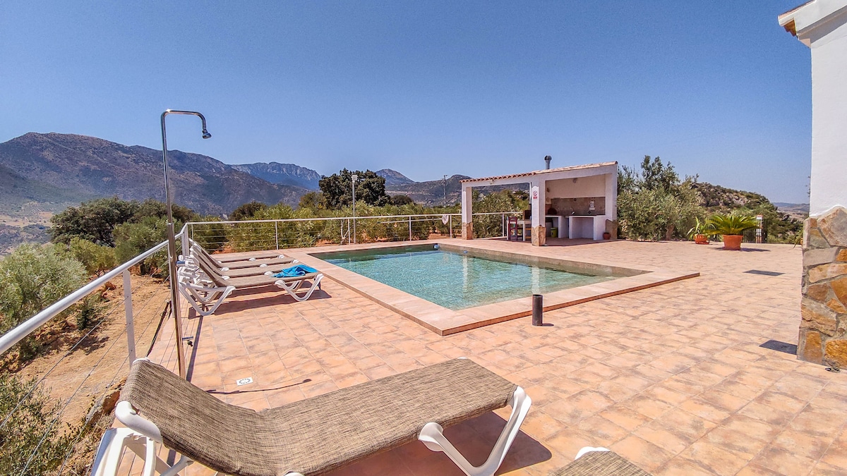 A private pool is set within a sunlit outdoor area, surrounded by tiled flooring and offering scenic views of the mountains. Sun loungers are positioned nearby, providing spaces for relaxation. An outdoor kitchen is visible, alongside potted plants that enhance the natural setting.
