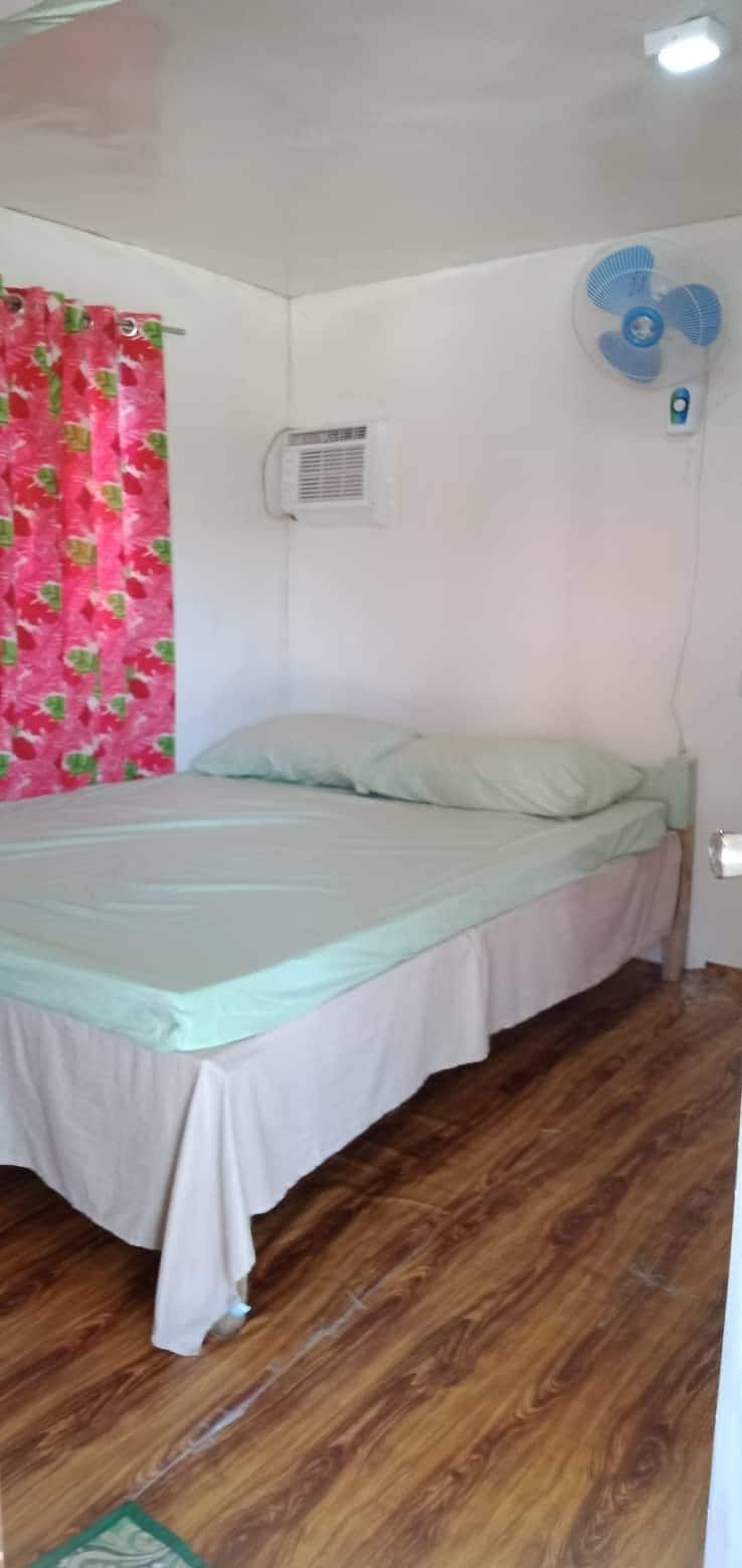 A simple bedroom features a spacious bed dressed in mint green linens. The room is accented by vibrant pink floral curtains. An air conditioning unit is mounted on the wall, and warm wooden flooring complements the bright, minimalist decor.