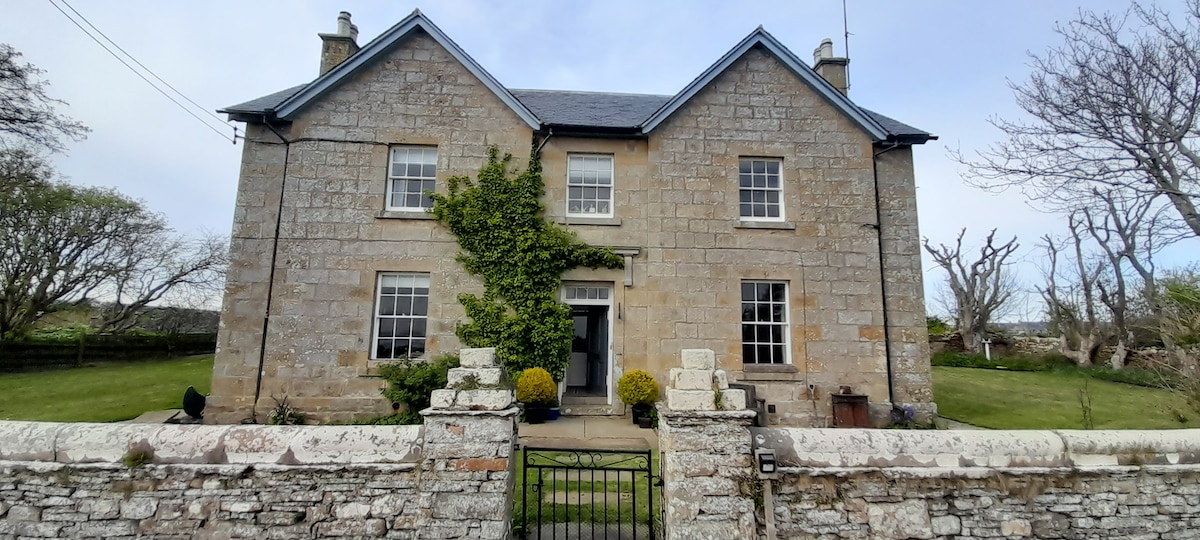 The historic exterior of the Old Manse is highlighted by its stone façade, characterized by symmetrical windows and a central doorway framed by greenery. A simple gate invites guests into the neatly maintained garden, which features various shrubs and trees, contributing to the serene ambiance.