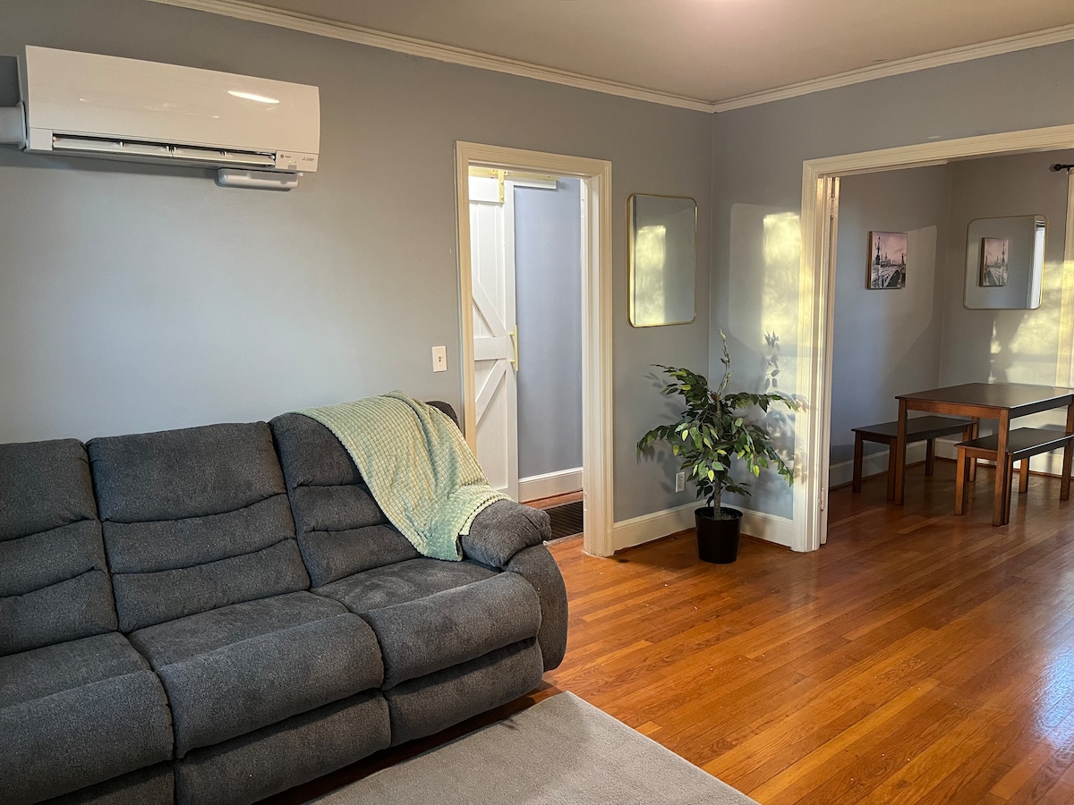 A cozy living area features a gray sofa with a light green throw blanket draped over one arm. A small table is visible nearby, along with a potted plant. Natural light enters through a nearby door, highlighting the warm wooden flooring and the simple decor.