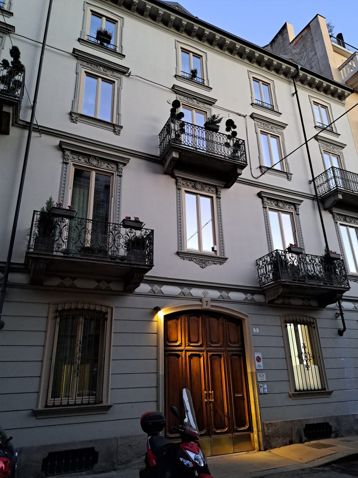 The building exterior showcases elegant architecture with intricate details. Large, framed windows are complemented by decorative balconies adorned with potted plants. A wooden entrance door serves as a focal point, while soft evening light reflects from the windows, creating a welcoming ambiance.