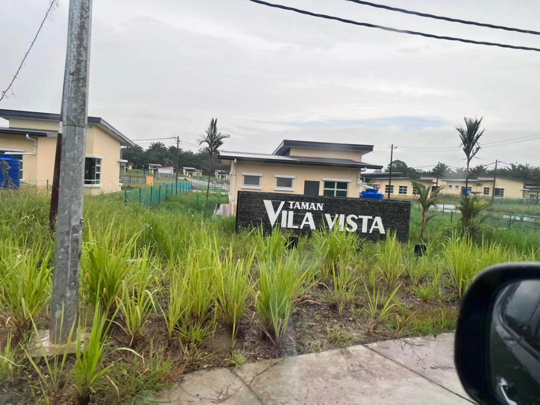 The entrance to Taman Vila Vista is displayed prominently, featuring a stylish sign set against a backdrop of greenery. Modern bungalow-style homes are visible in the distance, surrounded by well-maintained grass and palm plants, contributing to a natural ambiance.