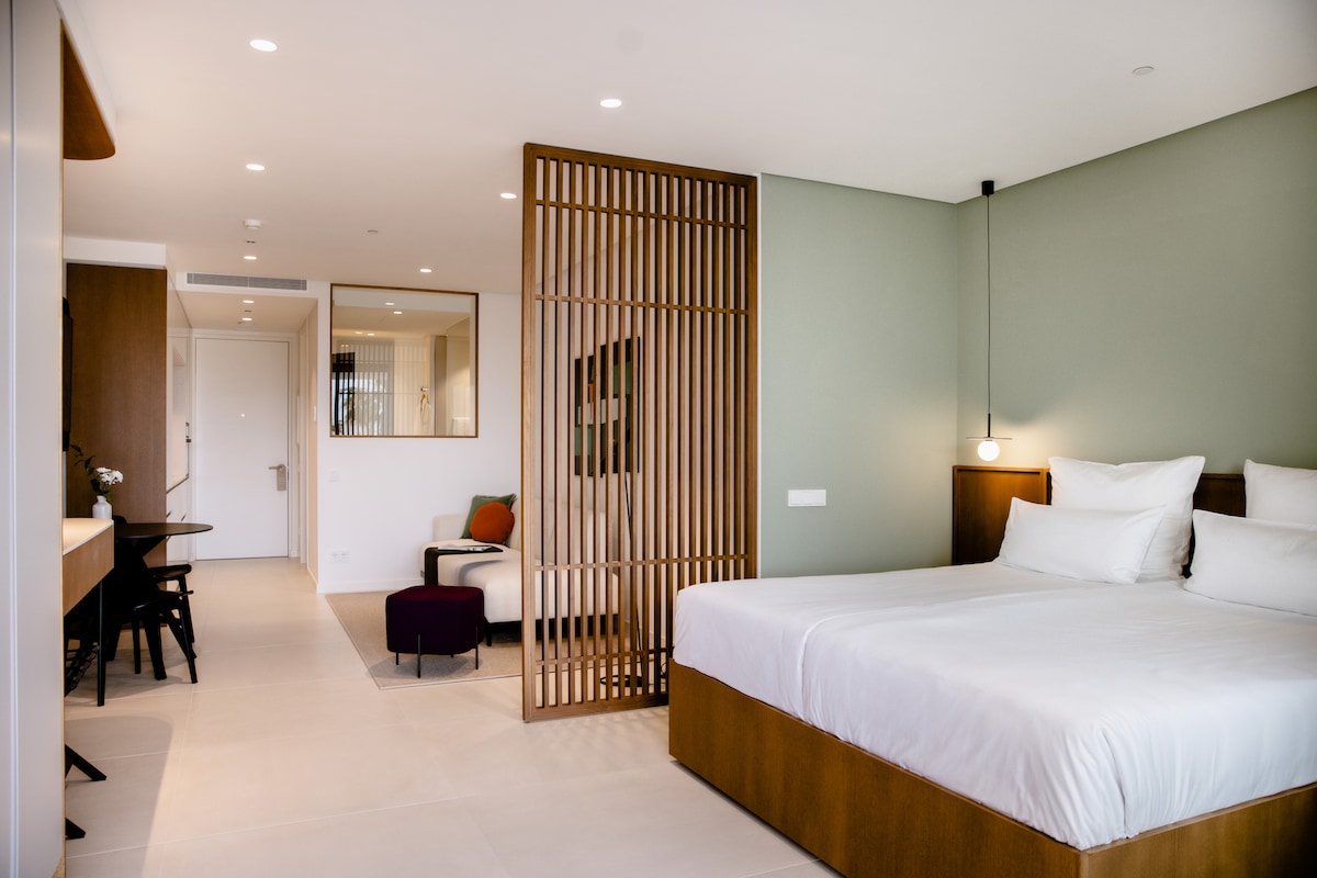 A bright studio space is featured, highlighting a spacious bed flanked by wooden accents. A modern sitting area is present, with a partition adding a sense of privacy. Soft lighting and neutral tones contribute to a calm environment, with a work desk positioned nearby.