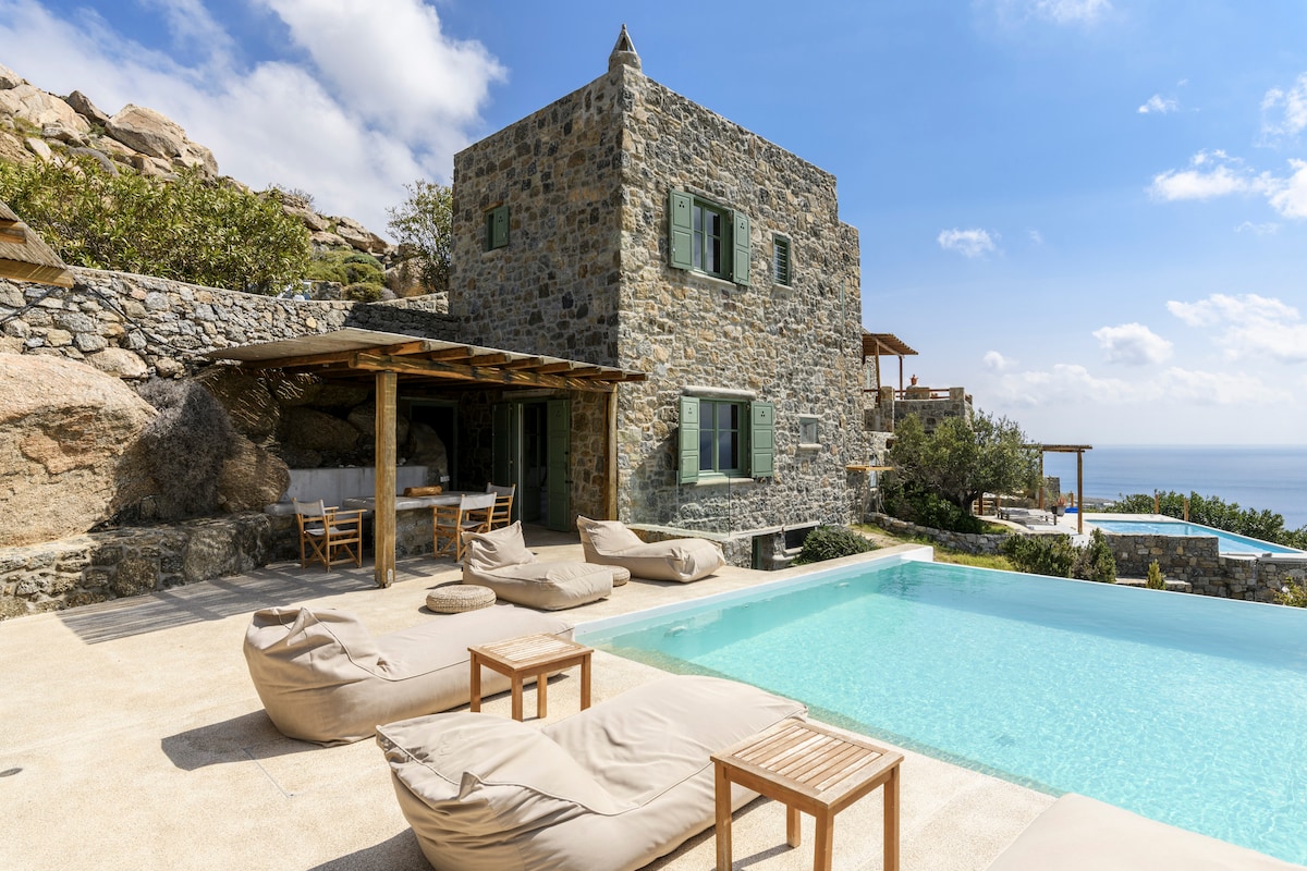 The exterior view of the villa showcases traditional dry stone walls complemented by green window shutters. A private infinity pool reflects the clear sky, surrounded by comfortable lounge seating and a wooden dining area. Natural vegetation enhances the tranquil outdoor atmosphere.