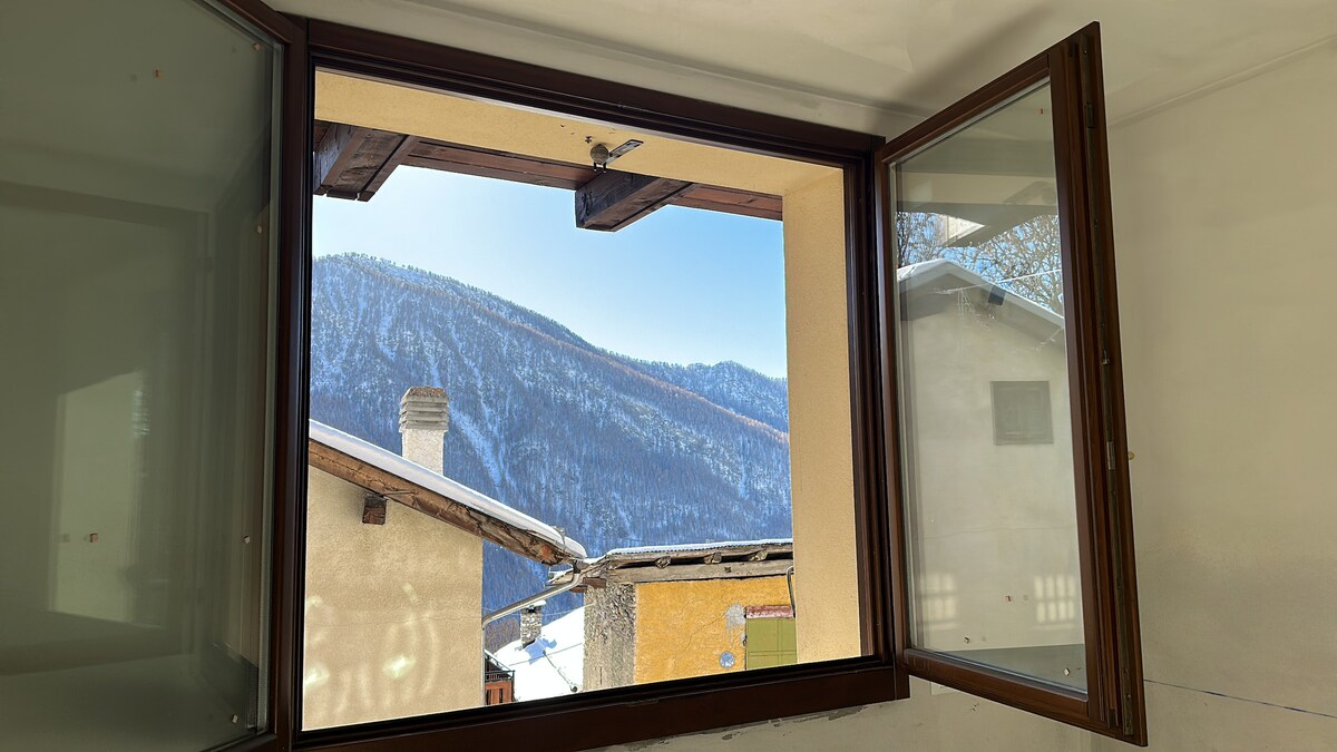 A view from the window reveals snow-covered mountains in the distance, framed by the building's wooden window design. Sunlight casts a gentle light over the landscape, enhancing the serene atmosphere of the surrounding area.