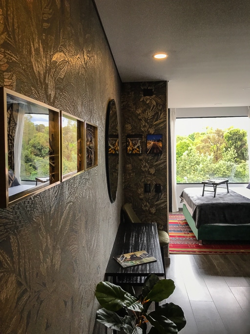 A modern loft space is depicted with a large window offering views of greenery. Decorative elements include framed photos and a circular mirror on the wall, alongside a low wooden table and a chair. A plush bed is positioned in the background, with soft natural light illuminating the room.