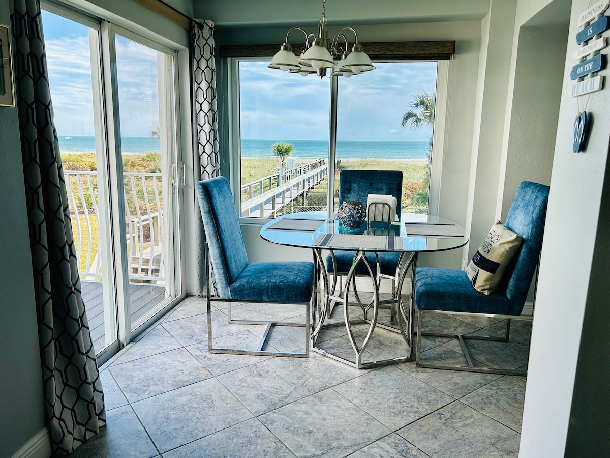 A dining area is illuminated by natural light through large windows offering ocean views. The space features a glass-top table surrounded by four blue upholstered chairs, with patterned curtains adding a touch of elegance. A view of the beach and boardwalk is visible outside.