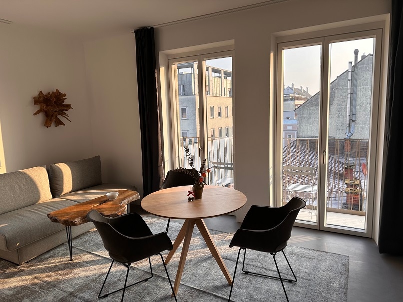 A bright living area is illuminated by natural light from large windows. A round wooden table is surrounded by two black chairs. A gray sofa sits against the wall, and a decorative wall piece adds visual interest.