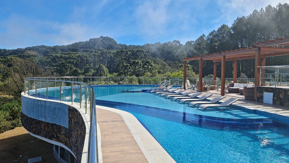 An expansive pool glistens under bright skies, surrounded by lush greenery. Lounge chairs are neatly arranged along the water's edge, inviting relaxation. A wooden pergola provides shaded seating nearby, enhancing the tranquil atmosphere of the resort.