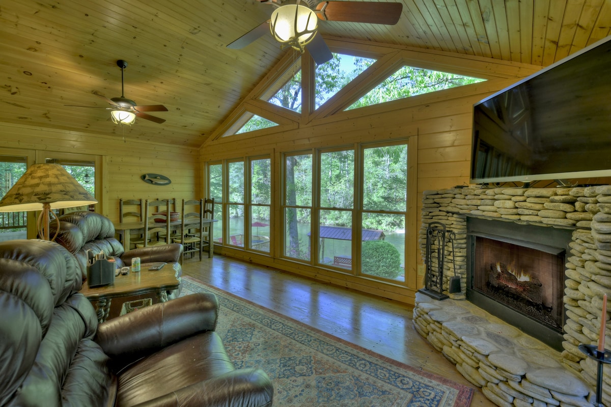 A spacious living area is defined by wooden paneling and large windows offering views of the outdoors. Comfortable seating is arranged around a stone fireplace, providing a cozy gathering space. Ceiling fans are present, contributing to a relaxed ambiance.