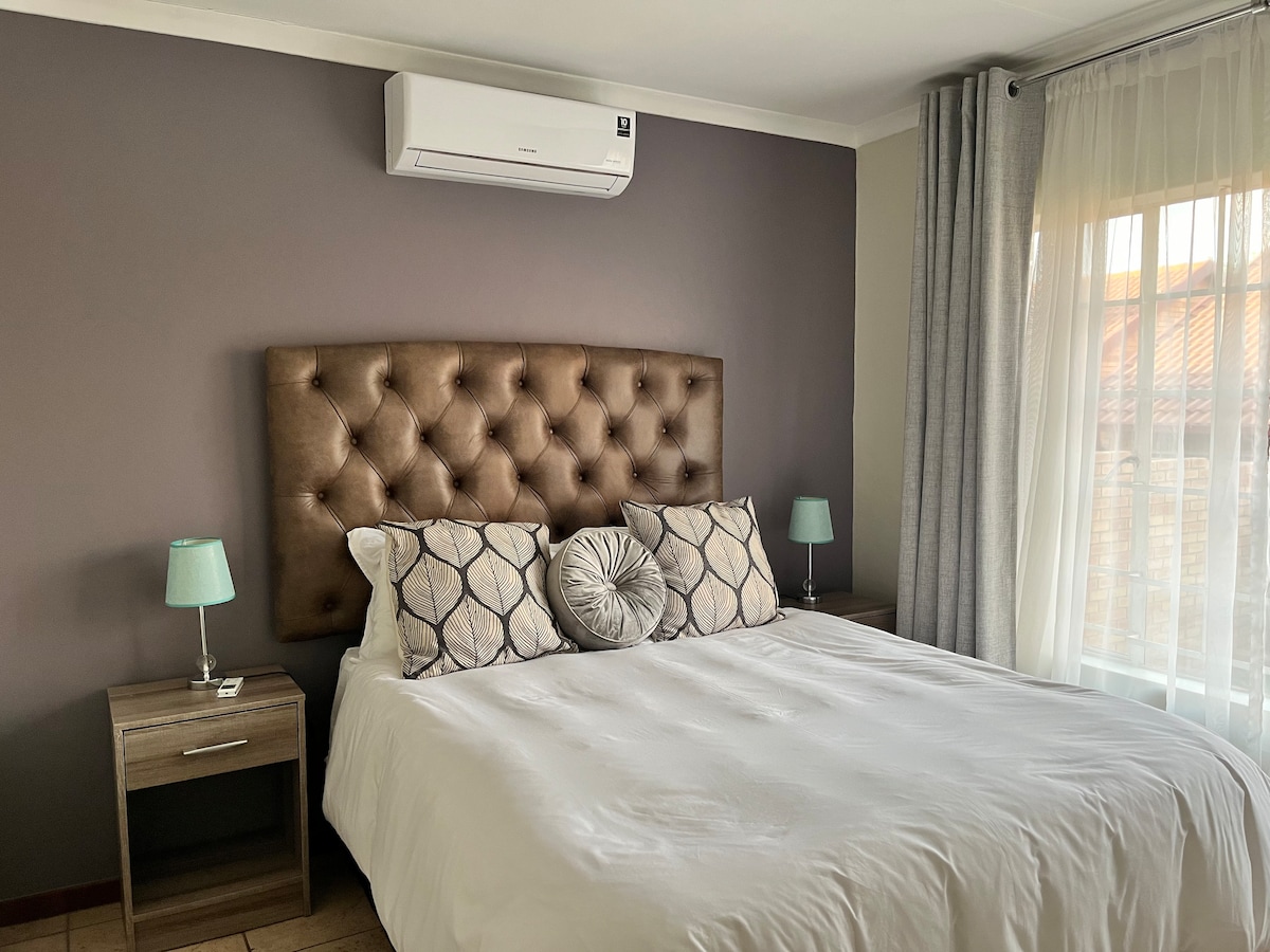 A well-appointed sleeping area features a spacious bed with a tufted headboard and decorative cushions. Light blue bedside lamps provide soft illumination. Floor-to-ceiling curtains frame the window, allowing natural light to enter the room, while a neutral color palette adds to the calming ambiance.