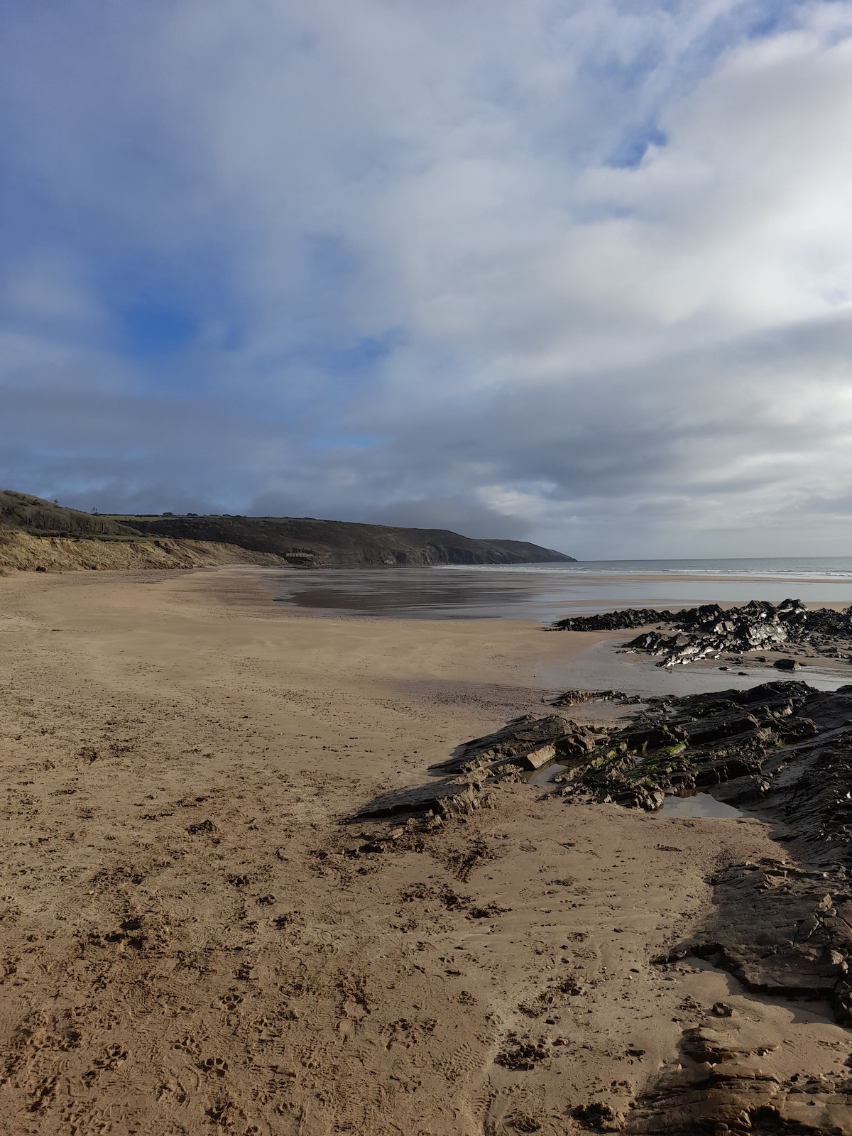 A sandy beach stretches into the distance, bordered by gentle waves lapping at the shore. Soft dunes and rocky formations are visible along the coastline, with a backdrop of low hills under a partly cloudy sky.