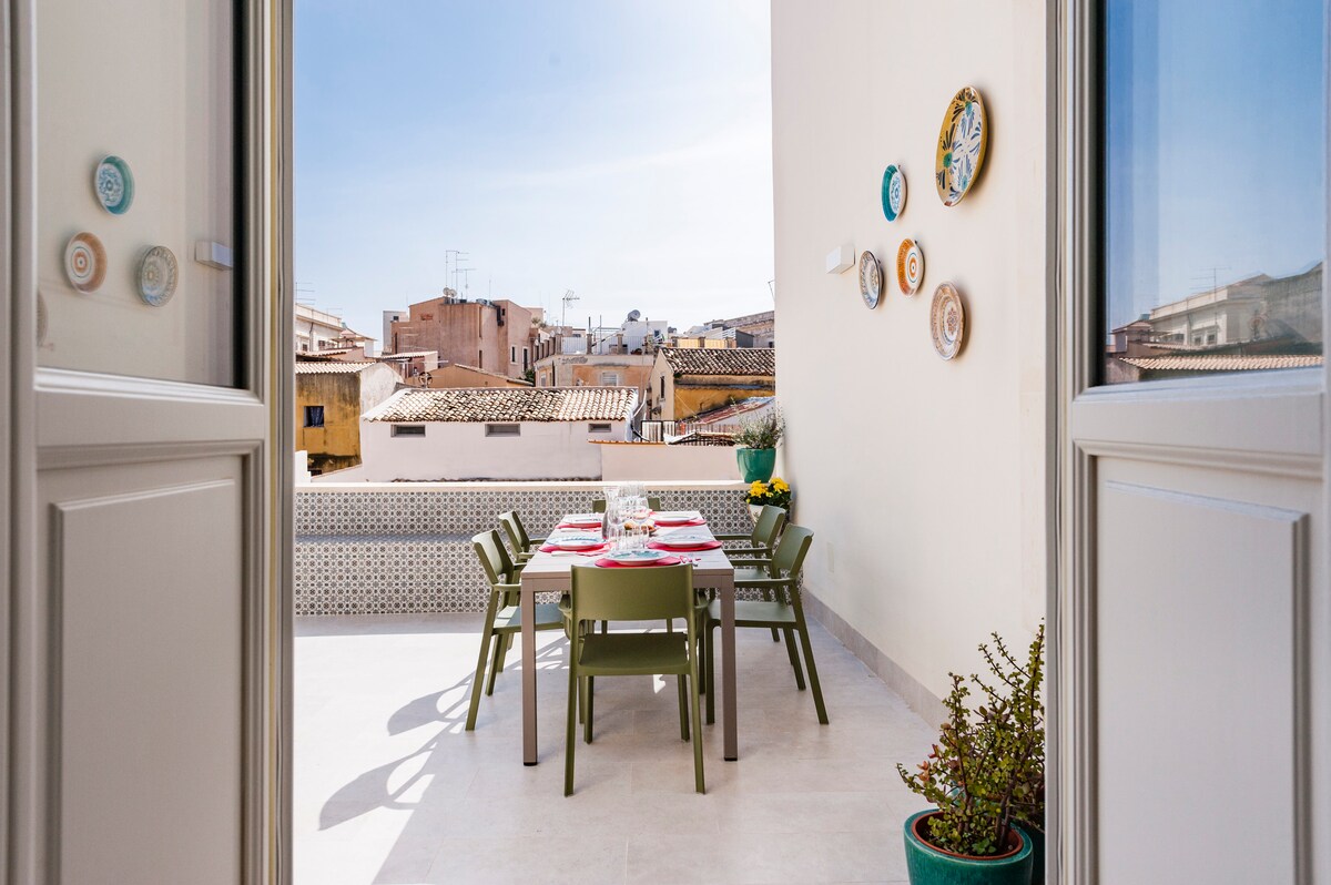 An outdoor terrace features a dining table set for meals, surrounded by green chairs. Decorative plates are mounted on the creamy wall, enhancing the space. A potted plant adds a touch of greenery. The view includes characteristic rooftops under a clear sky.