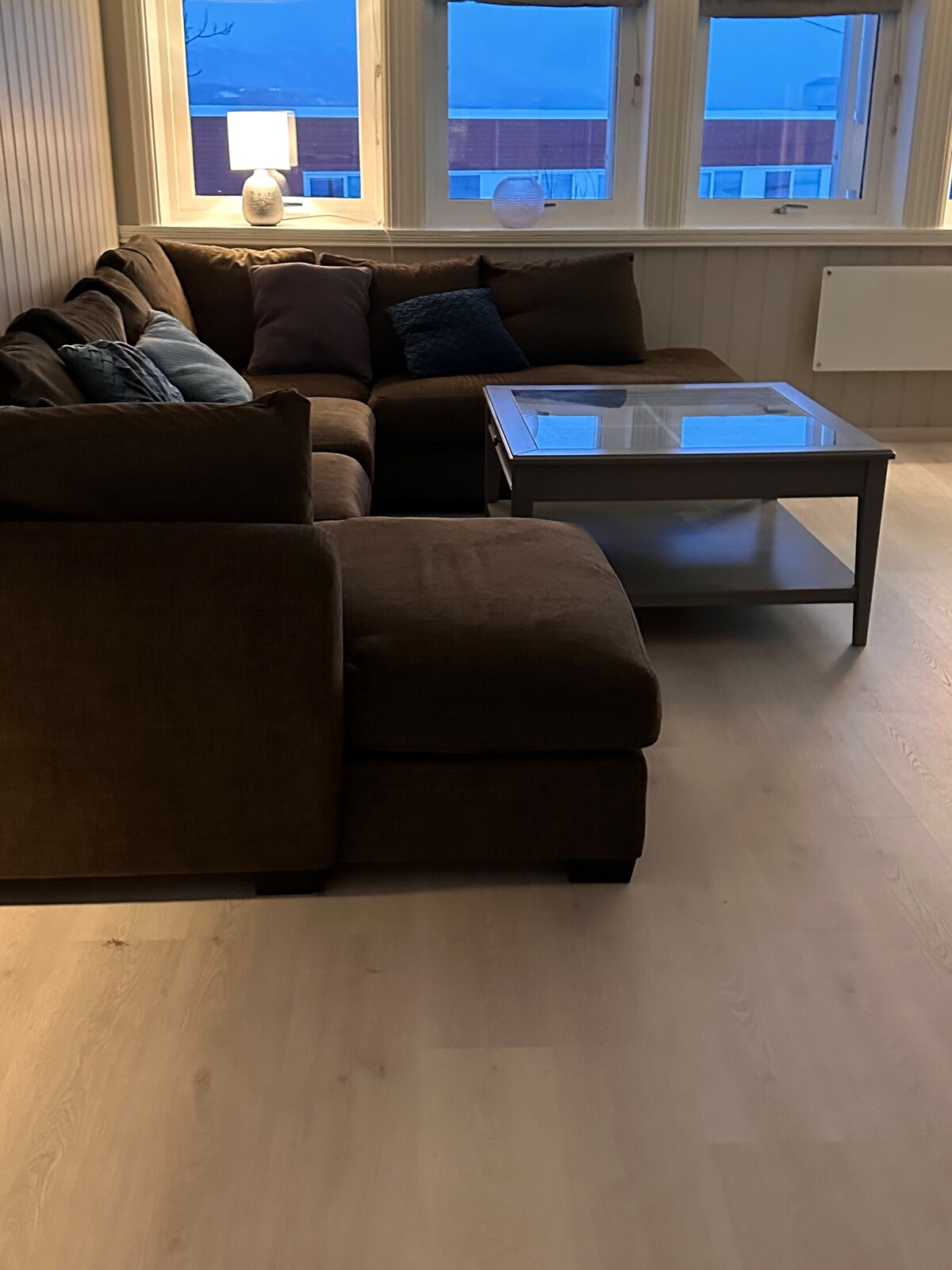 A cozy living area is featured, showcasing a brown sectional sofa adorned with various cushions in different shades. A glass coffee table sits in the center, positioned under soft lighting from a nearby lamp. Large windows allow natural light to fill the space.