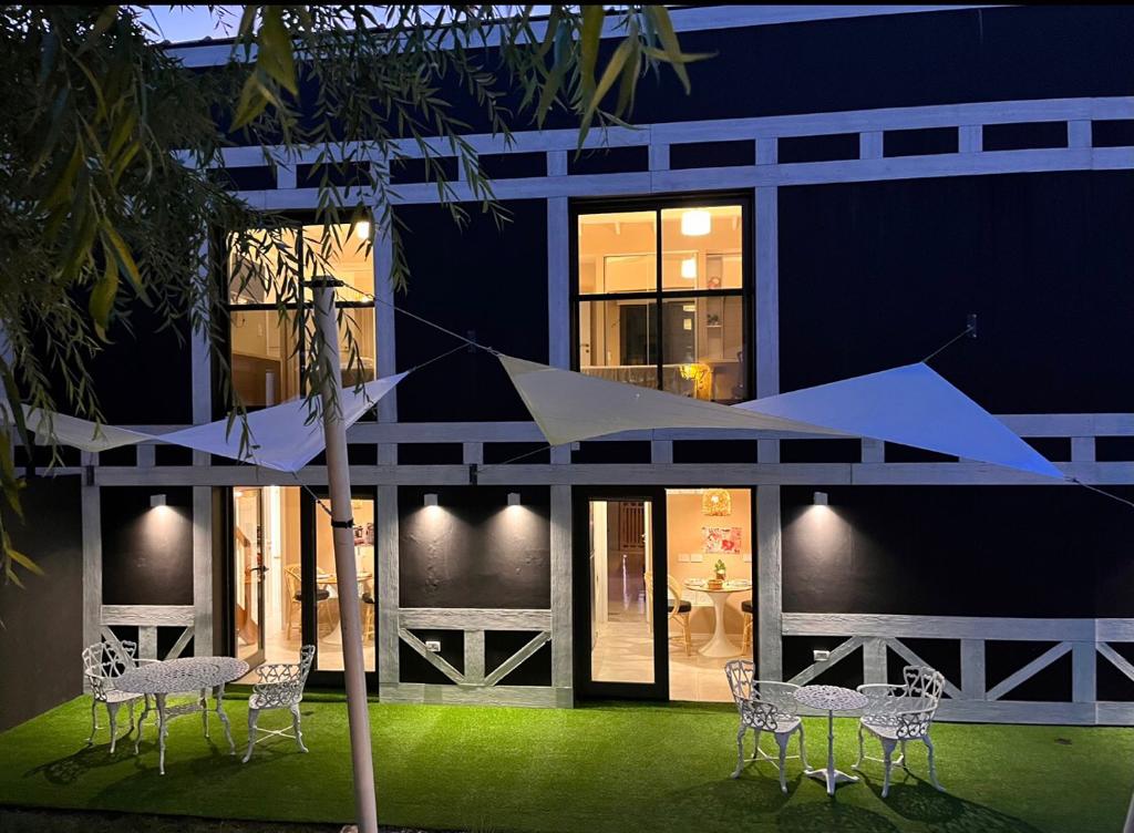 The exterior of the building is showcased at dusk, featuring modern glass doors and large windows. Two outdoor seating areas with white metal furniture sit on a grass-like surface, complemented by decorative canopies that provide shade.