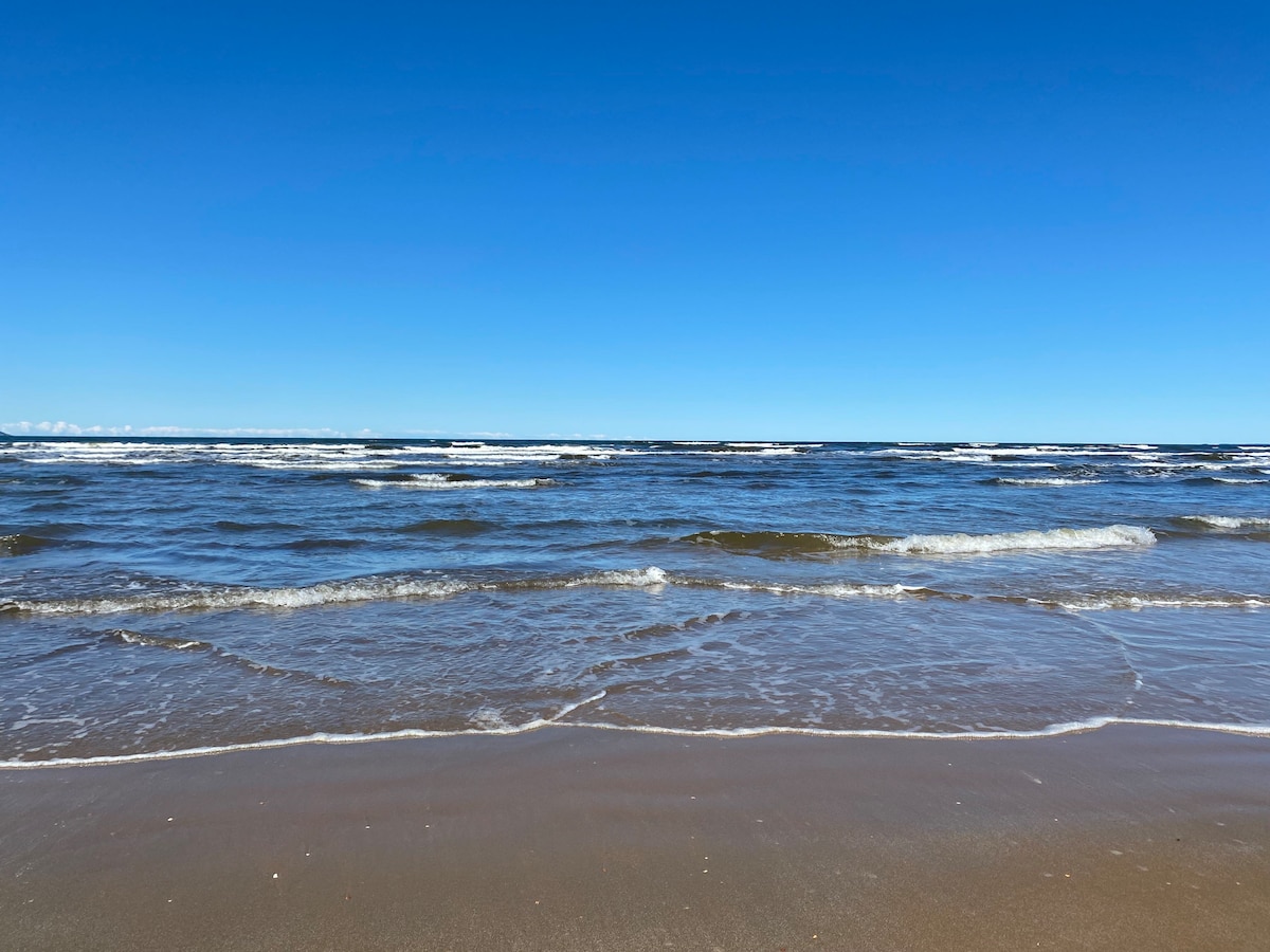 The image captures a clear view of the ocean, with gentle waves lapping against a sandy shore. The sky is bright blue, creating a serene backdrop for the rhythmic movement of the water. The scene suggests a lively beach atmosphere, welcoming various recreational activities.