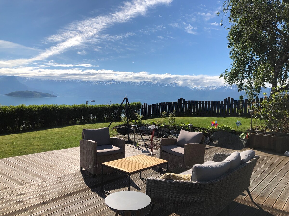 A well-furnished outdoor area features a simple wooden deck with lounge seating arranged around a coffee table. Scenic views of the fjord and mountains are visible in the background, with greenery framing the space. A fire pit is also present, enhancing the outdoor atmosphere.